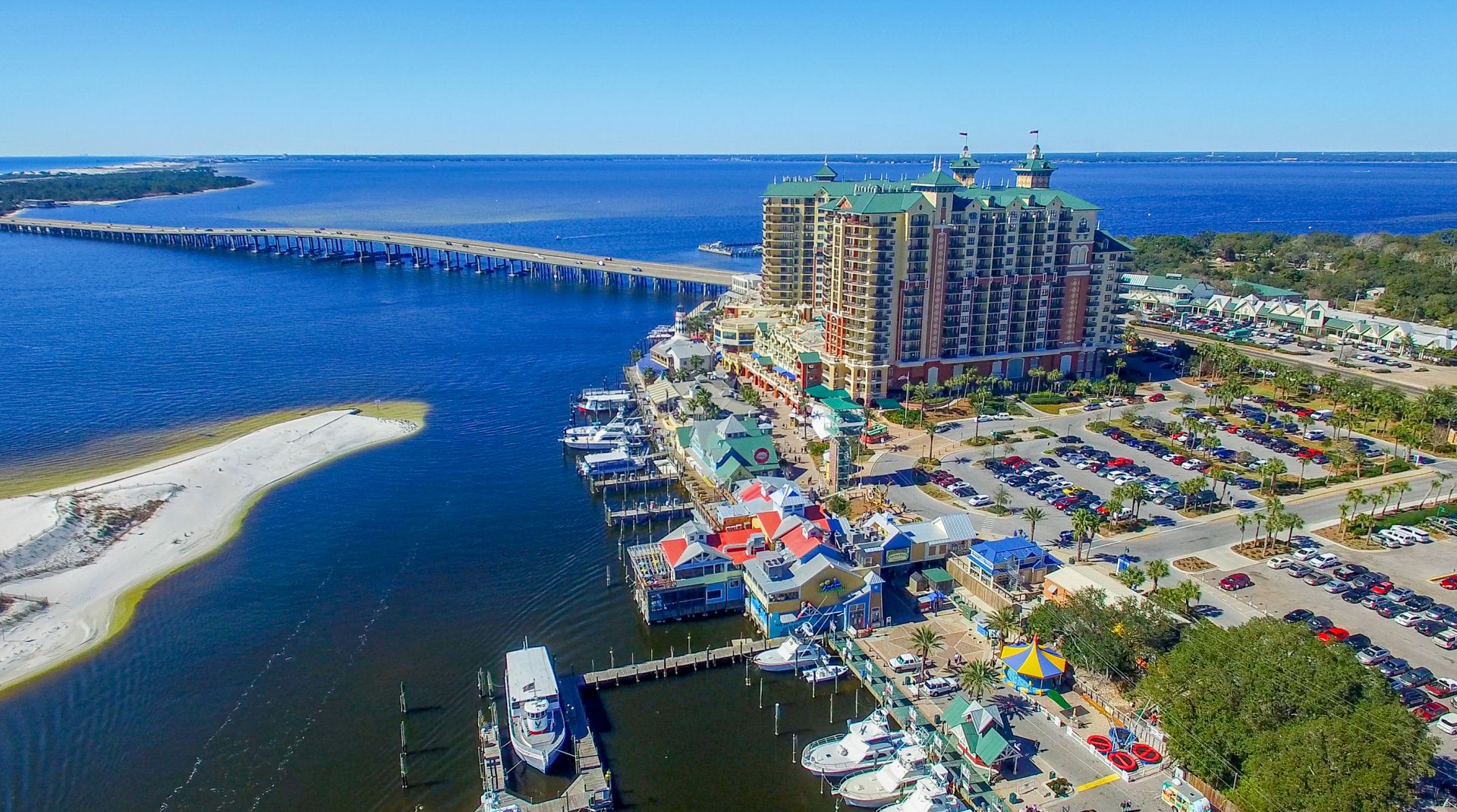 Aerial view of a coastal resort with a large multi-story hotel by the water, a long bridge crossing the bay, and a busy marina with boats and colorful waterfront structures on a sunny day.
