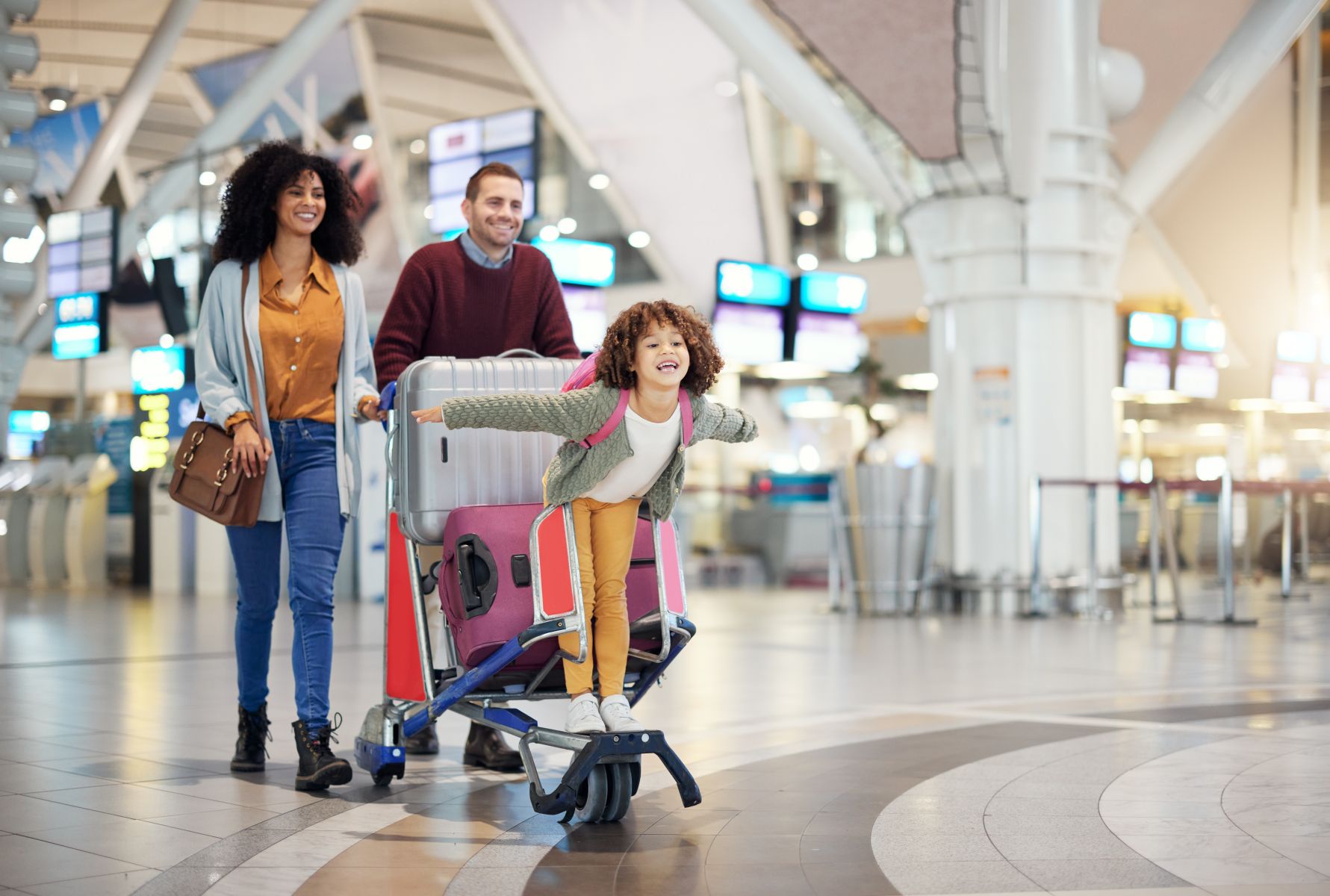 Family of three at an airport: mom and dad push a luggage cart with a large silver suitcase while their daughter stands on a pink suitcase and smiles with arms outstretched.