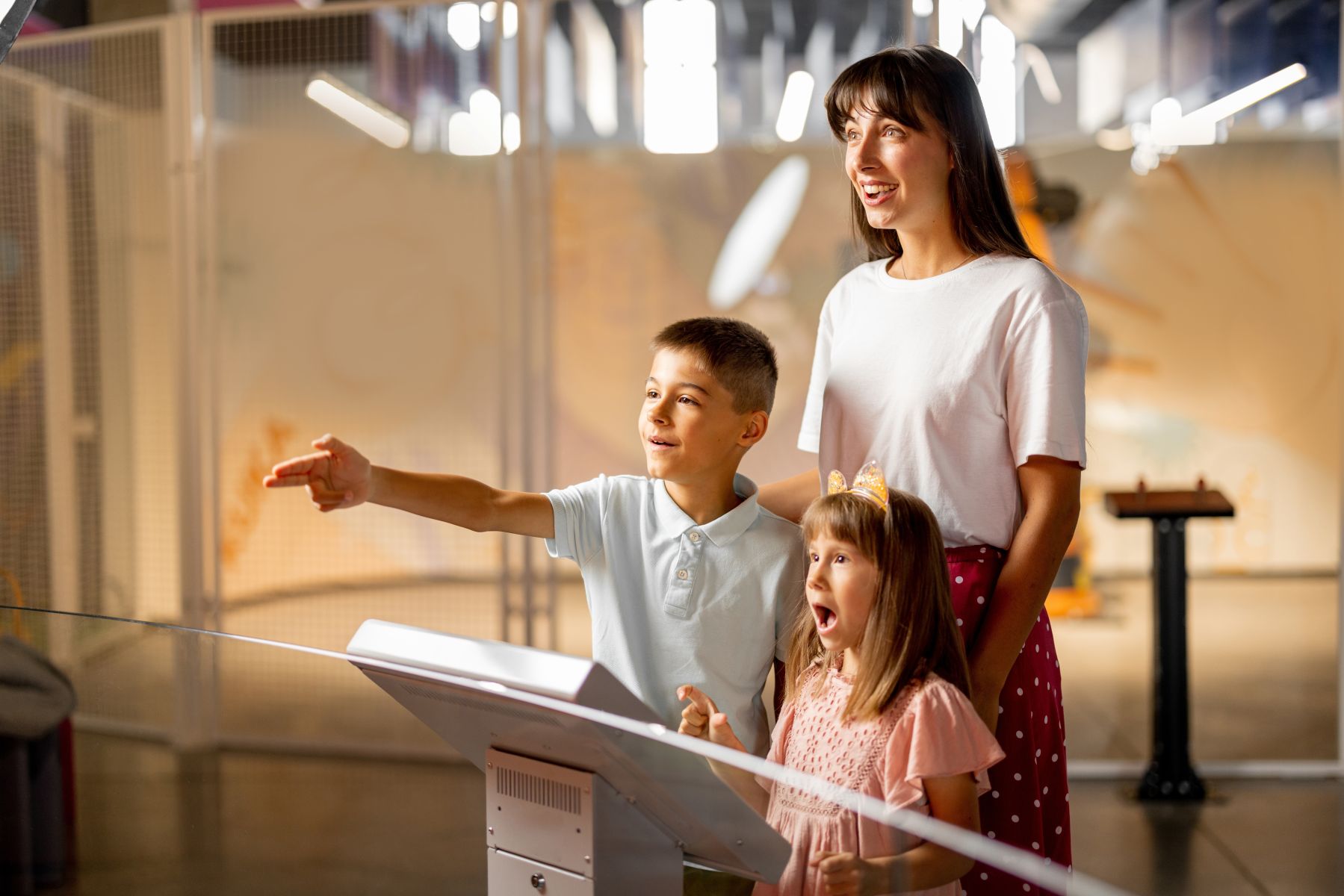 A woman with two children watches and points at an interactive museum exhibit with a touchscreen display front and center.