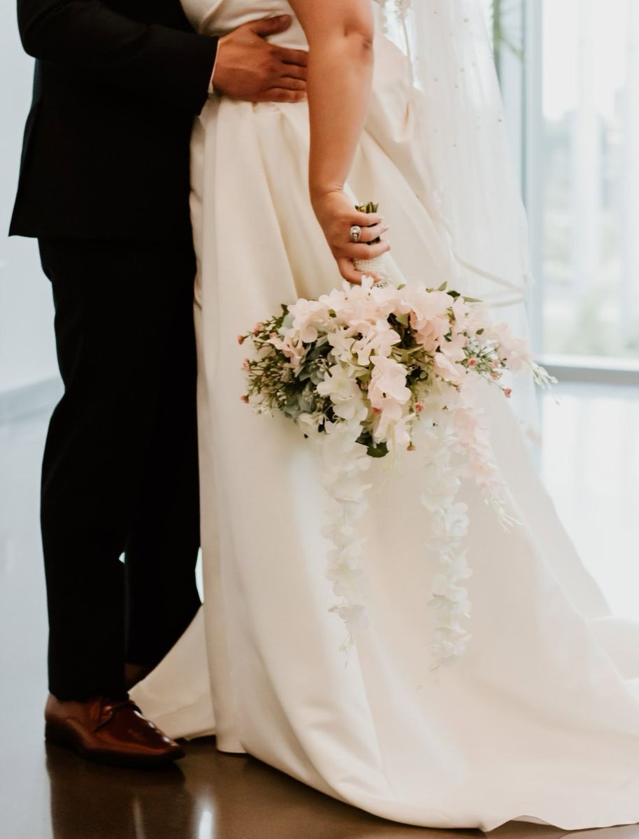 Bride in a white wedding dress holds a cascading bouquet of white and blush flowers as the groom stands beside her.