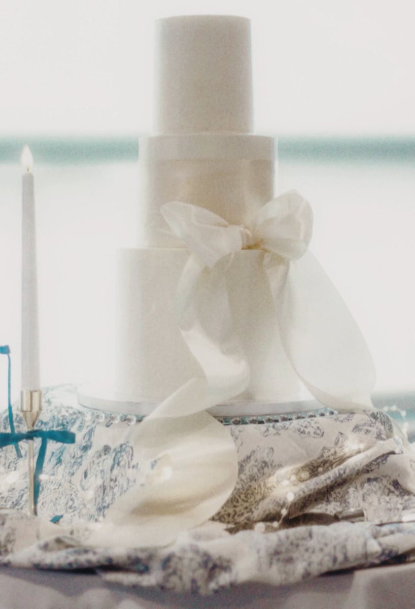 White three-tier wedding cake with a large satin bow, set on a decorative table near a lit candle.