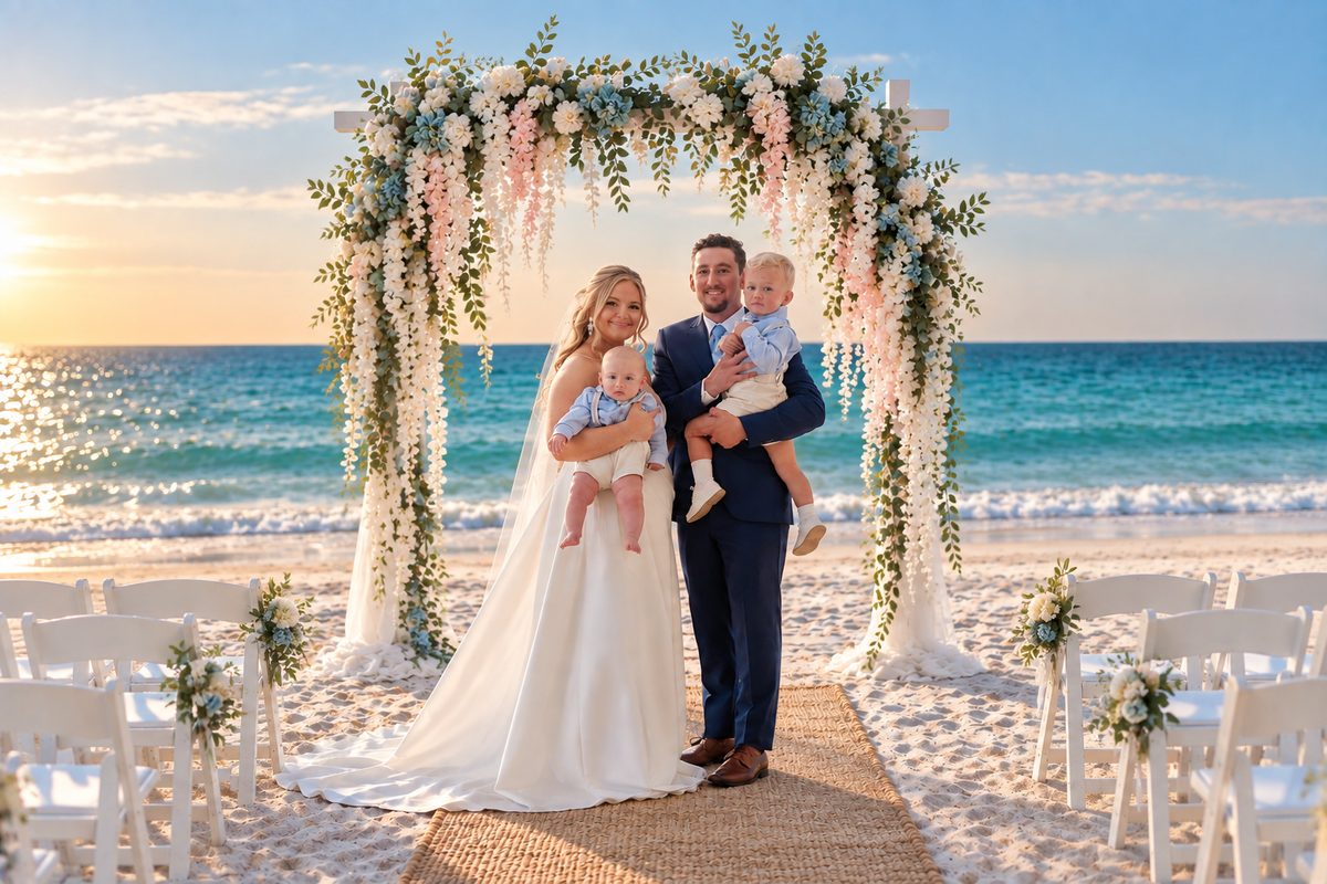 Bride and groom hold two babies under a floral arch on a sunny beach ceremony, ocean in the background on the sunset backdrop.