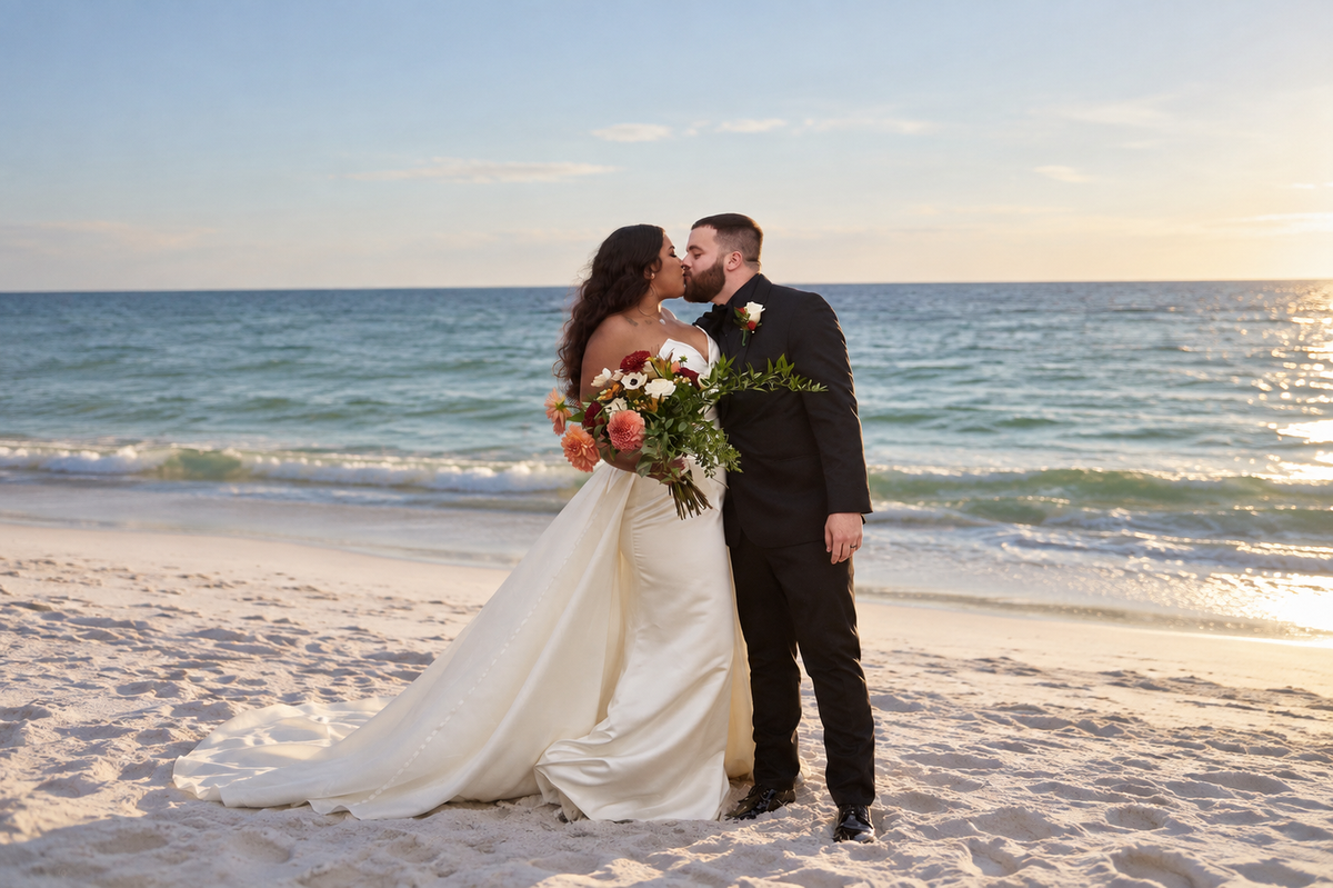 On a sandy beach, a bride in a white gown and a groom in a black suit kiss, holding a bouquet as waves and a sunset glow behind them.